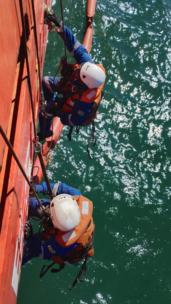 Dois t&eacute;cnicos suspensos em cordas ao lado do casco, vistos de cima, sobre o mar